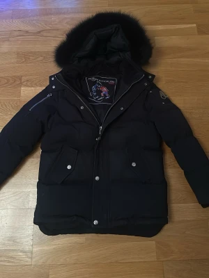 Svart  Moose Knuckles - Säljer en svart, vadderad pälsjacka från Moose Knuckles med fluffig pälskant på huvan. Jackan har flera fickor med lock och tryckknappar, samt dragkedja och tryckknappar framtill. Perfekt för kalla vinterdagar och riktigt snygg streetstil. Jackan passar dig som är mellan 145cm till 162cm