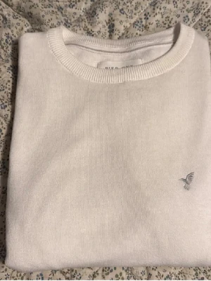 Vit stickad tröja pier one - Säljer en stilren vit stickad tröja med rund hals och ribbade muddar. Tröjan har en liten grå broderad fågel på bröstet som detalj. Perfekt för dig som gillar minimalistisk design och mjuka material.