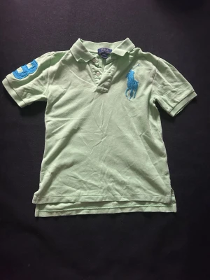 Ljusgrön piké från Polo Ralph Lauren - Snygg ljusgrön pikétröja från Polo Ralph Lauren med blå logga på bröstet och siffran 3 på ärmen. Klassisk krage och knappar framtill. Perfekt för en avslappnad men ändå stilren look. OBS! Barn storlek