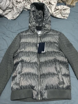 Grå Dior pufferjacka med huva - Snygg grå pufferjacka från Dior med ikoniskt monogrammönster över hela jackan. Jackan har stickade ärmar och muddar, dragkedja framtill och en stor huva. Materialet är en mix av glansigt tyg och stickat, vilket ger en unik look.