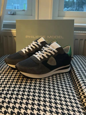 Philippe Model grå sneakers - Snygga sneakers från Philippe Model i grått och svart med detaljer i grönt och vitt. Ovandelen är i mesh och mocka, med vita skosnören och en vit sula. Klassisk låg modell som passar perfekt till streetwear och casual outfits. Helt nya aldrig använda. Pris kan diskuteras vid snabb affär!