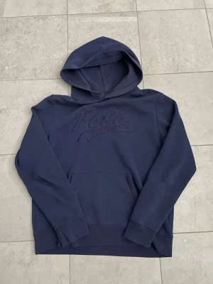 Mörkblå hoodie från Polo Ralph Lauren - Snygg mörkblå hoodie från Polo Ralph Lauren med broderad logga på bröstet. Klassisk känguruficka framtill och mjukt material som känns skönt mot huden. Perfekt för en avslappnad och stilren look. Huva med dragsko och ribbade muddar vid ärmar och nederkant.