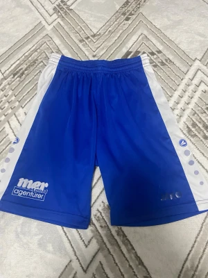 Blå sportshorts från JAKO - Snygga blå sportshorts från JAKO med vita sidopaneler och prickdetaljer. Shortsen har elastisk midja och tryckta loggor på framsidan. Perfekta för träning eller fotboll och tillverkade i lätt polyester för maximal komfort.