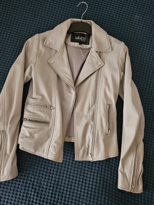 Ljusgrå skinnjacka från inbyM - Snygg ljusgrå skinnjacka från inbyM med klassisk bikerkrage, silverfärgade dragkedjor och tryckknappar. Jackan har flera fickor med dragkedja och en figurnära passform som ger en cool look. Perfekt för dig som vill ha en stilren och trendig jacka.