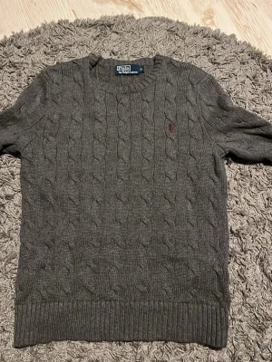  kabelstickad ralph lauren tröja - Kabelstickad grå tröja från Polo Ralph Lauren med rund halsringning och röd broderad logga på bröstet. Tröjan har ribbade muddar vid ärmslut och nederkant. Klassisk och tidlös design i mjukt material.