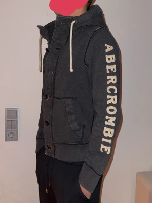 Mörkgrå hoodie från Abercrombie - Mörkgrå hoodie från Abercrombie med stora vita bokstäver längs ena ärmen. Tröjan har huva med vita dragsnören, knappar framtill och två fickor. Ribbstickade muddar vid ärmslut och nederkant. Perfekt för en avslappnad och cool stil.
