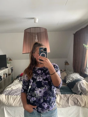 Mörkblå blommig topp med 3/4-ärm - Snygg mörkblå topp med lila och vita blommor. Modellen har 3/4-ärm och rund halsringning. 