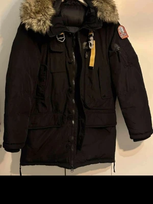 Svart Parajumpers parkas med päls - Snygg svart parkas från Parajumpers med avtagbar pälskant på huvan, flera fickor och ikonisk gul rem vid halsen. Jackan har patch på ärmen och robust dragkedja. Perfekt för kalla dagar och riktigt varm.