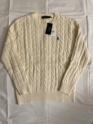 Vit kabelstickad tröja Polo Ralph Lauren - Kabelstickad vit tröja från Polo Ralph Lauren med rund halsringning och ribbade muddar. Klassisk marinblå logga broderad på bröstet. Tröjan är i mjukt material och har en tidlös design som passar till mycket.