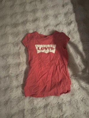 Rosa Levi's t-shirt med logga - Säljer en rosa t-shirt från Levi's med klassisk vit logga framtill. T-shirten har korta ärmar och rund halsringning. Tillverkad i mjuk bomull som är skön att bära. Perfekt för dig som gillar en enkel men snygg stil.