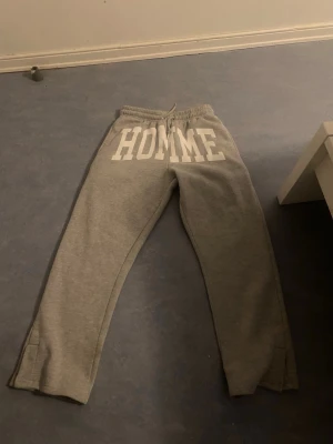 Gråa mjukisbyxor med HOMME-tryck - Säljer ett par gråa mjukisbyxor med stort vitt HOMME-tryck över framsidan. Byxorna har resår i midjan med dragsko och raka ben. Perfekta för chill dagar eller när du vill ha en avslappnad stil.