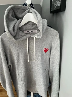 Grå hoodie från Comme des Garçons Play - Snygg grå hoodie från Comme des Garçons Play med klassisk huva och vita dragsnören. På bröstet sitter det ikoniska röda hjärtat med ögon. Hoodien har känguruficka och är gjord i mjukt bomullsmaterial. Perfekt för en chill och stilren look.
