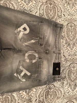 Grå jeans från Richmond - Säljer ett par grå jeans från Richmond med coolt spraymålat tryck på bakfickorna och slitna detaljer framtill. Jeansen har fem fickor, bälteshällor och en svart patch med logga vid linningen. Perfekt för dig som gillar streetstyle och unika plagg.