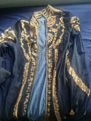 Svart broderad blus med guldmönster - En Unik marockansk abaya svart blus med transparent tyg och detaljerat guldigt broderi framtill och på ärmarna. Hög krage och puffiga långärmar ger en elegant look. Perfekt för dig som vill sticka ut med en statement topp.