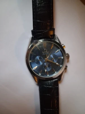 Omega Seamaster klocka svart läder - Stilren Omega Seamaster herrklocka med svart urtavla och tre mindre blåa subdials. Klockan har ett klassiskt silverfärgat boett och svart läderarmband med krokodilstruktur. Perfekt accessoar för dig som gillar tidlös design och exklusiv känsla. O.b.s batteribyte och sätta fast rem. Säljs billigt pga detta. Värdet är betydligt mer!