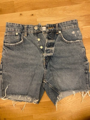 Högmidjade jeansshorts med rå kant - Säljer ett par klassiska blå denimshorts med hög midja och rå, fransig kant. Shortsen har fem fickor, knappar i gylfen och en avslappnad passform. 