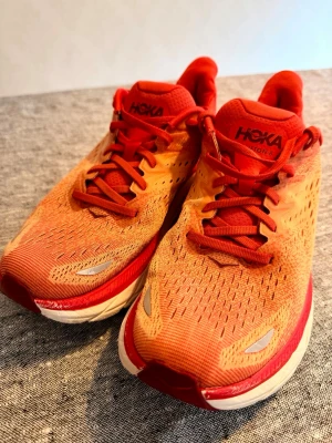 Hoka Clifton 8 löparsko, storlek 42 - Snygga Hoka Clifton 8 sneakers i en grymt fräsch orange och röd färgkombination. Ovandelen är i mesh för bra andningsförmåga och sulan är vit med röda detaljer. Perfekta för dig som gillar löpning eller vill ha riktigt bekväma skor till vardags. Reflekterande detaljer för extra synlighet.