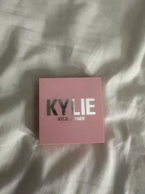 Kylie Jenner highlighter i guld - Highlighter från Kylie Jenner i en snygg rosa fyrkantig dosa med spegel. Pudret har en skimrande guldig nyans och ett mönstrat ytskikt. Perfekt för att ge glow till ansiktet och framhäva kindbenen.