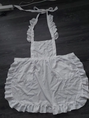 Vit volangförkläde cosplay - Sött vitt förkläde med volangkant och två stora fickor framtill, perfekt för cosplay eller maskerad. Klassisk modell med knytband i midjan och runt nacken. Tillverkat i ett lätt tyg, troligen polyester eller bomullsblandning.