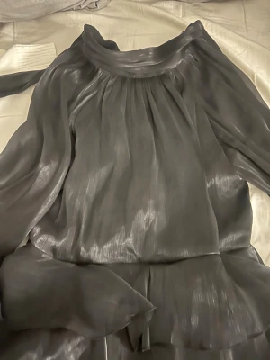 Grå omlottklänning i satin - Snygg grå omlottklänning i glansigt satinliknande material med volangdetaljer och lång ärm. Klänningen har en lös passform och ett elegant fall, perfekt för dig som gillar en stilren look med lite extra shine. Testat 1 gång köpt för ca 2 månader sen. 
