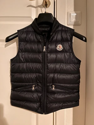Svart dunväst från Moncler - Storlek 12/152 Cm. Snygg svart dunväst från Moncler med dragkedja och två fickor framtill. Västen har en hög krage och passar perfekt till lite kyligare sommar/vår dagar. Jättebra kvalitet, inte några skador eller något liknande. 