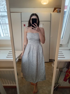 Ljusblå blommig långklänning - Långklänning med smockad överdel och tunna axelband. Klänningen är ljusblå med små blommönster och har en luftig, utsvängd form. Perfekt för varma dagar och har en feminin, avslappnad stil.