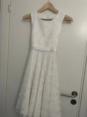 Fin klänning  - Elegant vit klänning med spetsmönster och ärmlös design. Klänningen har en vacker rosett baktill och ett band med blomdetalj i midjan. Perfekt för sommarens speciella tillfällen/bröllop.Dragkedja i ryggen och utsvängd kjol ger en klassisk siluett. Super fin,har en liten fläck som knappt syns.