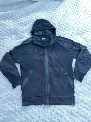 Svart zip hoodie CP Company - Säljer en svart hoodie från C.P. Company med dragkedja framtill och två snedställda fickor med dragkedja. Huvan har inbyggda glasögon och justerbara snören, vilket ger en unik och streetinspirerad look. Perfekt för dig som vill sticka ut med din stil.