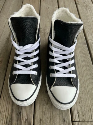 Svarta Converse All Star Highs - Säljer ett par svarta Converse All Star highs i läder med vit sula och tåhätta. Skorna har vit pälsfodring på insidan, vita skosnören och klassisk Converse-logga på sidan. Perfekta för dig som vill ha både stil och värme. Aldrig använda, bara blivit stående här! I originalkartong