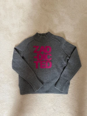 Grå stickad tröja från Zadig et Voltaire - Snygg grå stickad tröja med hög krage och breda ribbade muddar. Framtill står det 'ZADDICTED' i stora rosa bokstäver. Perfekt för dig som gillar statement-plagg och vill sticka ut lite extra. Materialet känns mjukt och värmande. Nypris 1600 kr! Älskar denna tröja 💕⭐️
