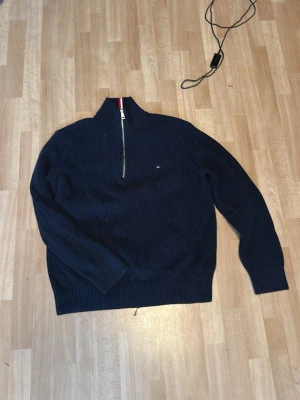 Marinblå halvzip från Tommy Hilfiger - Säljer en marinblå stickad tröja från Tommy Hilfiger i ekologisk bomull. Tröjan har halv dragkedja, hög krage med röd och vit randig detalj samt diskret logga på bröstet. Perfekt för dig som gillar klassisk och stilren design.