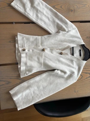 Vit kofta från Gina Tricot - Stilren vit kofta från Gina Tricot med v-ringning och tre stora bruna knappar framtill. Koftan har två fickor fram och ribbade muddar vid ärmslut och nederkant. Tillverkad i mjukt stickat material.💕