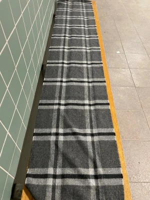 Grå halsduk med rutigt mönster - Snygg och lång halsduk i grått med klassiskt rutigt mönster i svart och vitt. Perfekt accessoar för att lyfta din outfit och hålla dig varm. Tjockt och mjukt material som känns skönt mot huden.