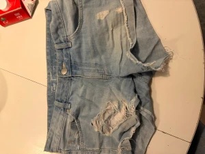 Ljusa blå jeansshorts med slitningar - Säljer ett par ljusa blå jeansshorts med råa kanter och slitna detaljer framtill. Shortsen har fem fickor, bälteshällor och knappgylf. Perfekta för sommaren och ger en avslappnad vibe. Midjemåttet rakt över är 43 cm💕