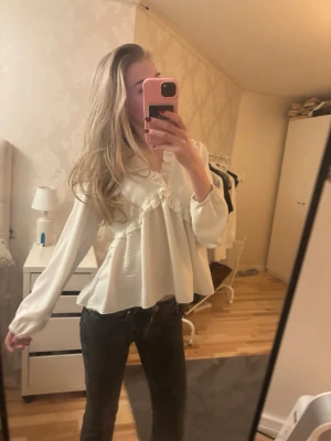 Vit blus med volangdetaljer - Säljer en vit blus med volangdetaljer över bröstet och v-ringning. Blusen har en luftig passform och långa ärmar med resår vid ärmslut. Perfekt för dig som gillar en romantisk och trendig stil. Storleken är one sized men sitter som storlek S. 😊