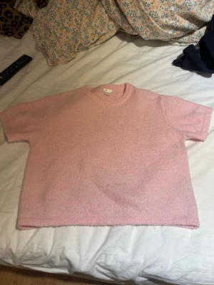 Ljusrosa stickad t-shirt - Söt ljusrosa stickad t-shirt med korta ärmar och rund hals. Tröjan har en boxig passform och är gjord i ett mjukt, fluffigt material som känns skönt mot huden. Perfekt för dig som gillar en avslappnad men ändå trendig stil.