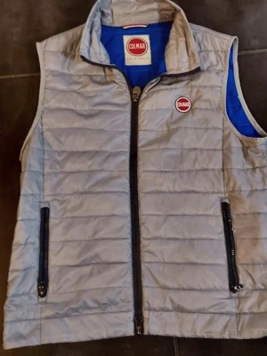 Ljusgrå dunväst från Colmar - Snygg ljusgrå dunväst från Colmar med blått foder och två svarta dragkedjefickor framtill. Västen har ståkrage och Colmar-logga på bröstet. Perfekt för lager-på-lager och enkel att matcha med olika outfits.