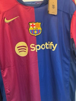 Barcelona matchtröja Nike blå/röd - Säljer en officiell FC Barcelona matchtröja från Nike i klassiska blå och röda färger. Tröjan har klubbmärke, Spotify-logga och Nike Swoosh framtill samt UNHCR-logga bak. Tillverkad i lätt och ventilerande material, perfekt för fotboll eller träning.