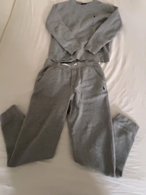Grått loungewear-set från Polo Ralph Lauren - Mysigt grått loungewear-set från Polo Ralph Lauren med sweatshirt och matchande byxor. Båda plaggen har den klassiska broderade loggan på bröstet och höften. Tillverkat i mjukt bomullsmaterial med avslappnad passform och rund halsringning.