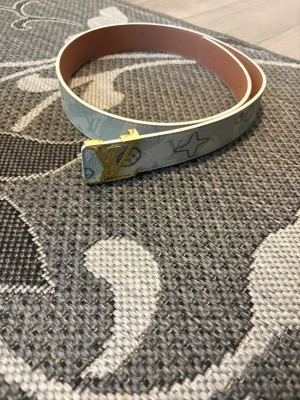 Louis Vuitton vitt bälte med guldspänne - Snyggt vitt bälte från Louis Vuitton med ikoniskt LV-mönster och ett stilrent guldigt LV-spänne. Bältet har en smal design och är tillverkat i skinn med tryckt logotyp och detaljer i ljusblått och grått. Perfekt accessoar för att lyfta din outfit.