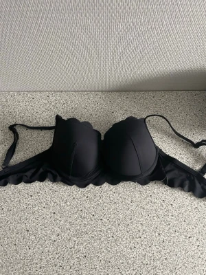 Svart vadderad bikini  - Snygg svart bikini med vadderade kupor och scallop-detaljer längs kanterna. A kupa i storlek 36, Justerbara axelband för perfekt passform. Kuporna ser större ut än vad dom är.