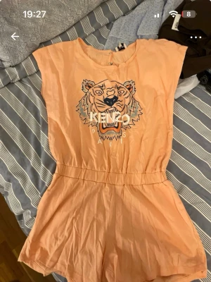 Persikofärgad Kenzo jumpsuit - Säljer en persikofärgad jumpsuit från Kenzo med ikoniskt tigertryck och logga på bröstet. Kortärmad modell med resår i midjan och rund halsringning. Materialet känns lätt och luftigt, perfekt för varma dagar. Supersnygg och trendig design! Helt ny och oanvänd! Så fin på sommarn dessutom. Barn storlek 