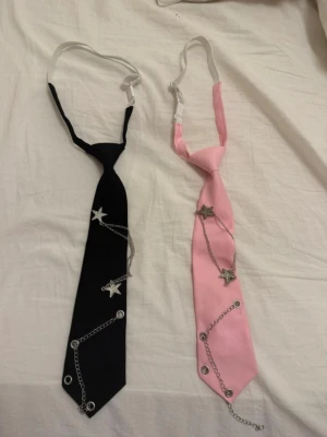 Två slipsar med kedjor och stjärnor - Två smala slipsar, en svart och en rosa, dekorerade med silverfärgade kedjor och stjärnformade detaljer. Slipsarna har öglor och metallringar som ger en alternativ och trendig look. Perfekt accessoar för att sticka ut och addera edge till din outfit.