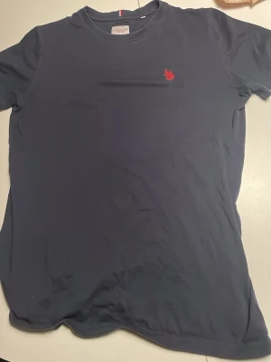 Mörkblå t-shirt från U.S. Polo Assn. - Mörkblå t-shirt från U.S. Polo Assn. med klassisk rund halsringning och röd broderad logga på bröstet. Tillverkad i mjuk bomull för en skön känsla. Enkel och stilren design som passar till det mesta.