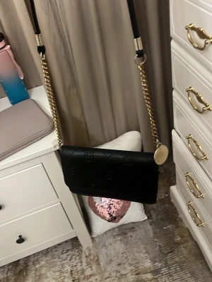 Svart axelväska med guldkedja - Snygg svart axelväska i quiltat mönster med guldfärgade kedjedetaljer och justerbar axelrem. Väskan har ett elegant lock och diskreta loggor i materialet. Perfekt för dig som vill ha en stilren och trendig accessoar.har några skador 