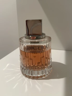 Jimmy Choo parfym med glasflaska - Elegant parfym från Jimmy Choo i en rund, genomskinlig glasflaska med räfflat mönster och silverfärgat lock. Doften har en varm, bärnstensfärgad vätska som syns genom flaskan. Perfekt för dig som gillar lyxiga och stilrena parfymer.