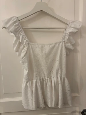 Vit volangblus med broderi - Supersöt vit blus med volangdetaljer på axlarna och broderade hålmönster. Toppen har en luftig passform, knytning i ryggen och är ärmlös. Perfekt för varma dagar och ger en romantisk vibe till din outfit.