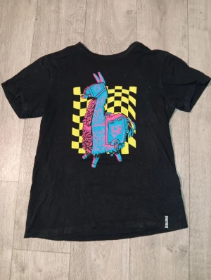 Svart Fortnite t-shirt med lama - Cool svart t-shirt från Fortnite med en färgglad grafisk print av en blå och rosa lama mot en gul-rutig bakgrund på framsidan. T-shirten har rund hals och korta ärmar. Perfekt för dig som gillar gaming och vill sticka ut med en unik design.