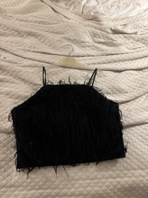 Svart fjädrig festtopp Gina Tricot - Säljer en svart festtopp från Gina Tricot med tunna axelband och fjädrig textur som ger en cool och trendig look. Toppen är croppad och perfekt för dig som vill sticka ut lite extra. 