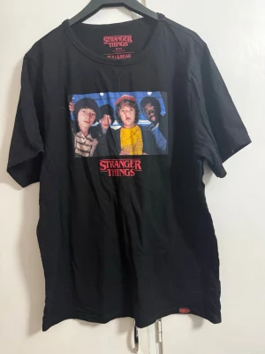 Stranger Things svart t-shirt - Cool svart t-shirt från Pull&Bear med Stranger Things-tryck på bröstet och en färgglad bild från serien. Klassisk passform och rund halsringning, perfekt för fans av serien som vill ha en casual look.